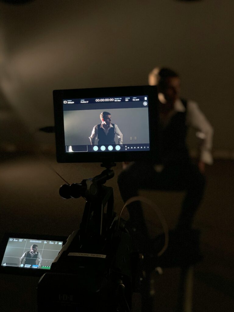 A well-dressed man poses in a studio with professional camera equipment shooting a performance.